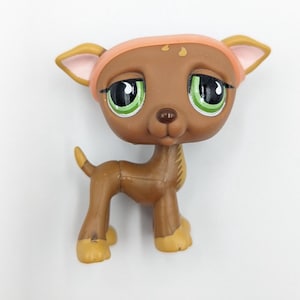 May include: A brown plastic deer figurine with large green eyes, wearing a pink headband. The deer has tan ears and feet. The figurine is a collectible toy.