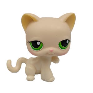 Puede incluir: Figura de gato de color beige claro con grandes ojos verdes y nariz rosa. El gato tiene una cola curvada y está de pie. Las orejas son de un tono ligeramente más oscuro de beige.