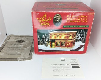 May include: A red box with a white and gold label that reads "A Wonderful Holiday." The box contains a miniature replica of a building with the text "The Bailey Bros. Building & Loan of Bedford Falls." The building is lit up with small lights and has a snowy scene around it.