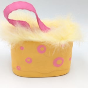 Könnte beinhalten: Ein kleiner, gelber Behälter mit rosa Tupfen und einem rosa Griff. Der obere Rand ist mit flauschigen, hellgelben Federn besetzt. Der Behälter ist wahrscheinlich ein Dekorationsartikel oder eine kleine Handtasche.