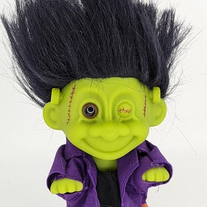 Puede incluir: Muñeco troll verde con pelo negro, un ojo y puntadas en la cara. Lleva una chaqueta morada y pantalones naranjas.