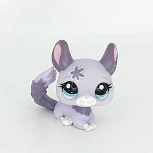 Könnte beinhalten: Kleine, sammelbare Chinchilla-Figur in Lavendel- und Grautönen. Das Spielzeug hat große, ausdrucksstarke Augen mit blauen Akzenten, eine weiße Schnauze und eine dekorative Blume auf dem Kopf. Das Chinchilla sitzt.