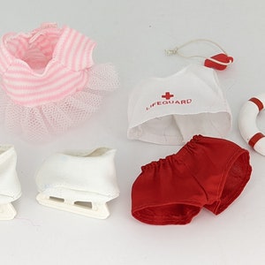 Puede incluir: Un conjunto de ropa para muñecas que incluye un vestido de rayas rosa y blancas con falda de tul, una camiseta de salvavidas blanca con una cruz roja y el texto "LIFEGUARD", pantalones cortos rojos, patines de hielo blancos y un salvavidas blanco con rayas rojas.