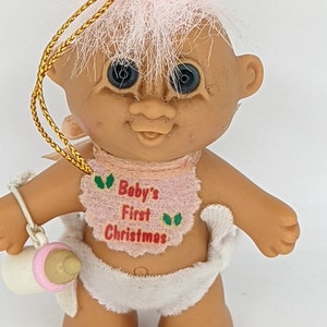 Puede incluir: Un adorno de troll de color rosa y blanco con un babero rosa que dice "Baby's First Christmas". La muñeca sostiene un biberón blanco y rosa.