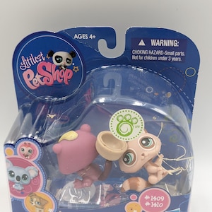 Puede incluir: Un juego de juguetes de Littlest Pet Shop en un paquete de plástico transparente. El juego incluye dos pequeñas figuras de animales, una rosa y otra beige, con accesorios. El embalaje tiene el texto "Littlest Pet Shop", "Ages 4+" y una advertencia de peligro de asfixia.