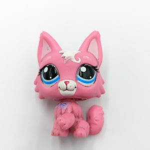 Puede incluir: Un gato de juguete de plástico rosa con ojos negros grandes, detalles azules y reflejos blancos. El gato tiene una pequeña nariz rosa y una mancha blanca en la cabeza. Está sentado erguido con las patas delanteras. Es un objeto de colección.