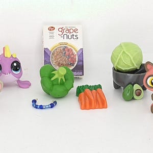 May include: A collection of colorful toy figurines and miniature food items. Includes a gray mouse, a purple and yellow creature, a box of Grape-Nuts cereal, a green pepper, a blue bracelet, orange carrots, a cabbage, avocados, and other animal figures.