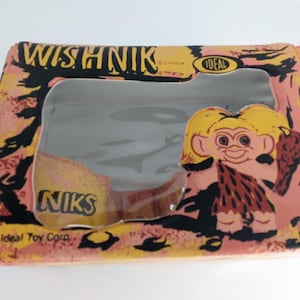 Puede incluir: Una caja de cartón rosa y amarilla con una ventana de plástico transparente. La caja tiene las palabras "Wishnik" e "Ideal Toy Corp." impresas en ella. La ventana muestra un troll de dibujos animados con cabello amarillo y un taparrabos marrón.