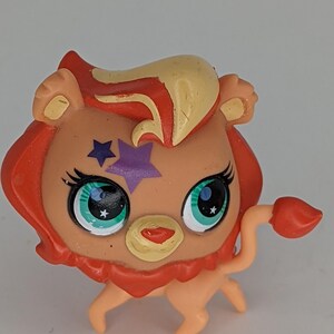Puede incluir: Una pequeña figura de león naranja y amarilla con una estrella morada en la frente y ojos verdes. La figura es parte de una colección de juguetes.