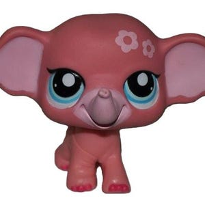 Op de afbeelding: Een roze olifantenbeeldje met grote oren en grote blauwe ogen. De speelgoedolifant heeft witte bloemaccenten op zijn hoofd en roze accenten op zijn oren en voeten. De olifant staat op vier poten.