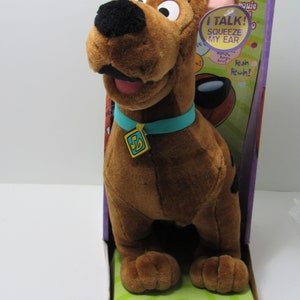 Puede incluir: Un peluche de Scooby-Doo en una caja. El perro marrón tiene una nariz negra, un collar turquesa con una etiqueta dorada y la boca abierta. La caja dice "I TALK! SQUEEZE MY EAR" y "SCOOBY-DOO! Talking Scooby-Doo".