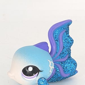Könnte beinhalten: Fischfigur mit blau-weißem Farbverlauf, lila Details und glitzernden blauen Flossen. Der Fisch hat ein großes Auge mit schwarzer Pupille und eine weiße Markierung an der Seite. Ein Sammlerstück.