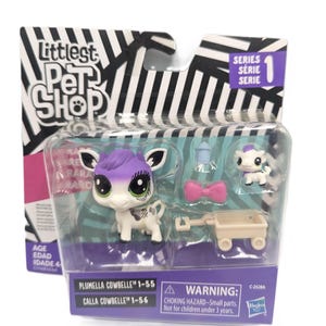 Puede incluir: Un set de juguete Littlest Pet Shop Plumella Cowbelle en su empaque original. Incluye una figura de vaca blanca y morada, una figura más pequeña, un lazo rosa, un biberón azul y un carrito beige. El empaque presenta rayas blancas y negras.