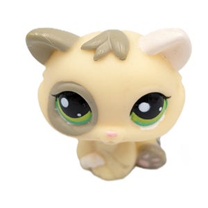 Puede incluir: Una figura de gato beige y gris con grandes ojos negros y detalles verdes. El gato de juguete tiene una nariz rosa y orejas pequeñas y detalladas. La figura es un juguete coleccionable.