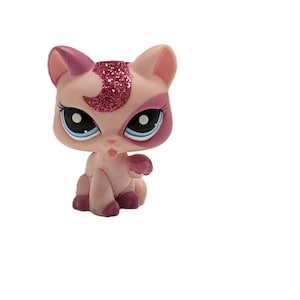 Könnte beinhalten: Hellrosa Katzenfigur mit dunkelrosa Akzenten. Die Katze hat große blaue Augen, ein rosa Glitzerdesign auf dem Kopf und sitzt. Die Figur ist ein Sammlerspielzeug.
