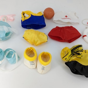 Puede incluir: Una colección de ropa y accesorios de muñecas en miniatura, que incluye un traje de baño azul con ribete de encaje, pantalones cortos amarillos y rojos, una camiseta de salvavidas y un salvavidas en miniatura. También hay zapatos y una pelota.