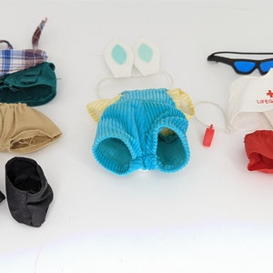 Puede incluir: Un conjunto de ropa y accesorios para muñecas, que incluye una caña de pescar con un pez, un bolso verde, pantalones cortos color beige, zapatos negros, orejas de conejo, un conjunto de pana azul, gafas de sol, un gorro de salvavidas, un salvavidas y pantalones cortos rojos.