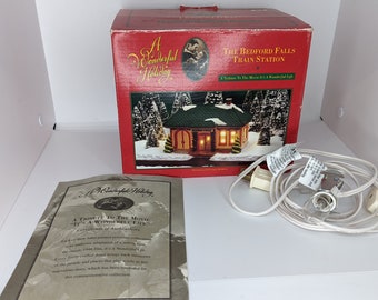 May include: A white box with a red lid and a picture of a small, lit, wooden house with a green roof. The box is labeled "The Bedford Falls Train Station". The box is sitting on a white surface with a white cord and plug.