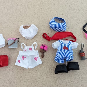 Puede incluir: Una colección de ropa y accesorios para muñecas, que incluye un atuendo de salvavidas, un atuendo de pintor, un atuendo de marinero y un traje de baño rosa. Los accesorios incluyen un salvavidas, un cubo de pintura, un pincel, una linterna y una visera.