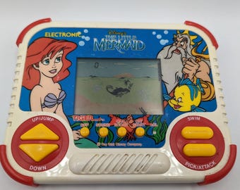 Op de afbeelding: Een vintage draagbaar elektronisch spel met personages uit Disney's De Kleine Zeemeermin. Het spel heeft een blauwe achtergrond met Ariel, Koning Triton en Botje. Het heeft rode en gele knoppen en een klein scherm met een spelscène.