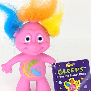 May include: A pink troll doll with blue, orange, and yellow hair. The doll has a crescent moon and a spaceship painted on its chest. The doll is holding a tag that says "GLEEPS From the Planet Bliss" with a smaller image of a troll doll on it.