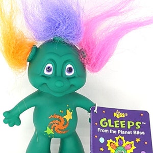 May include: A green troll doll with orange, purple, and pink hair. The doll has a galaxy design on its chest and is holding a tag that reads "Russ® GleePs™ From the Planet Bliss".