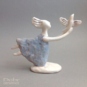 May include: A ceramic sculpture of a woman in a blue dress holding out a white bird. The woman has flowing hair and is in a flying pose. The sculpture is on a white base.