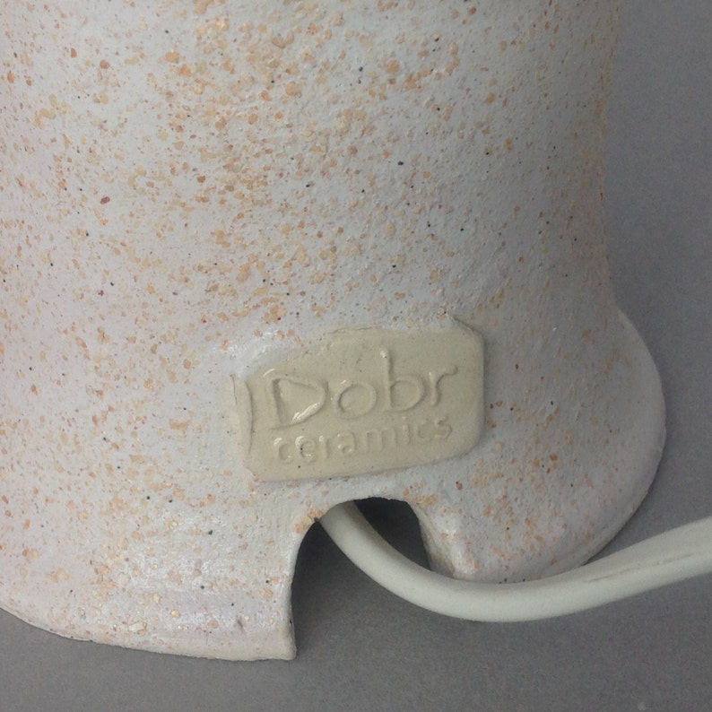 Op de afbeelding: Een witte keramische lampenvoet met een gespikkelde textuur. De voet heeft een kabelgat en een label met de tekst "Dobr ceramics".