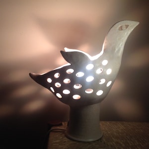 Op de afbeelding: Een witte keramische lamp in de vorm van een vogel met een licht dat door de gaten in het lichaam schijnt. De lamp heeft een ronde voet.