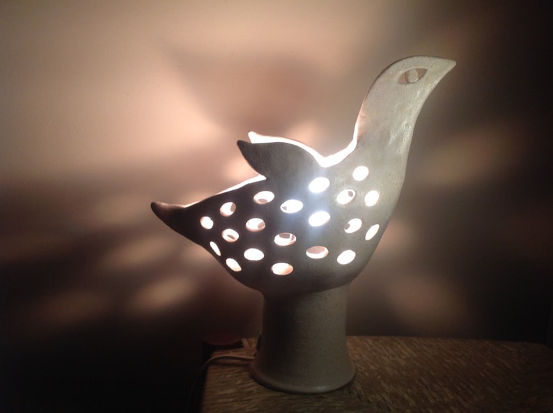 Op de afbeelding: Een witte keramische lamp in de vorm van een vogel met een licht dat door de gaten in het lichaam schijnt. De lamp heeft een ronde voet.