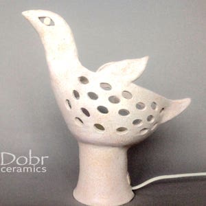 Op de afbeelding: Witte keramische vogellamp met een uitgesneden ontwerp en een snoer. De lamp staat op een grijs oppervlak. "Dobr ceramics" staat in zwarte tekst op het oppervlak.