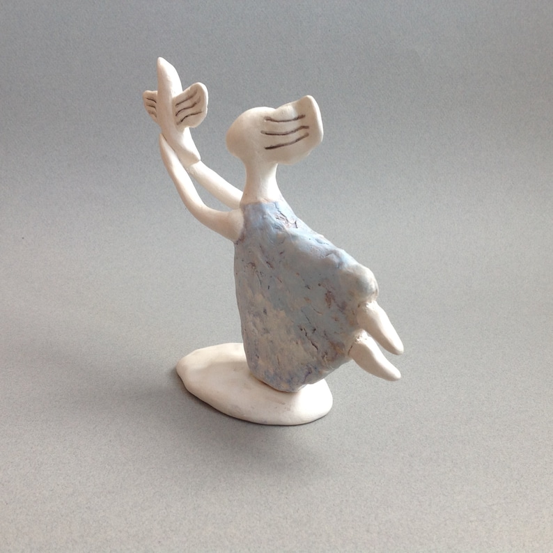 May include: A white ceramic figurine of a person holding a bird in their outstretched hand. The person is wearing a light blue dress and has a white base.