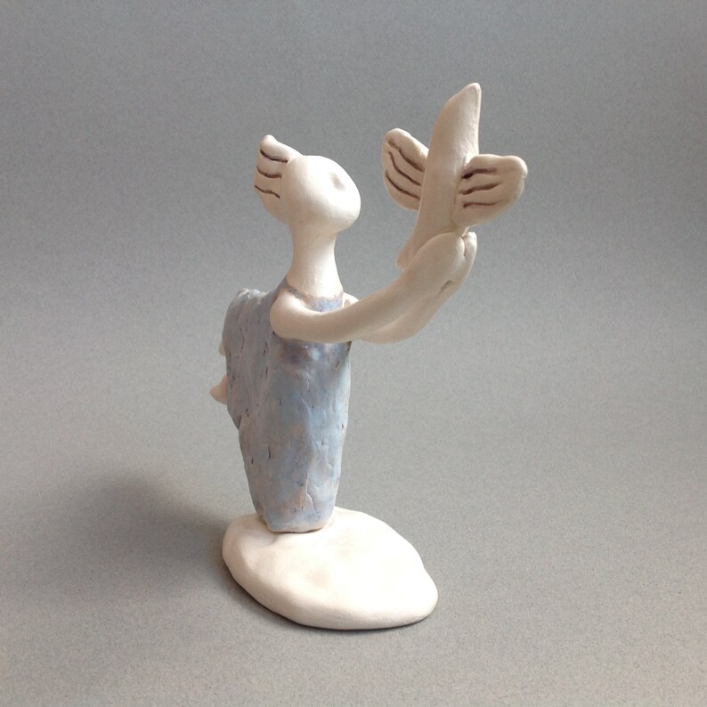 May include: A ceramic figurine of a person holding a bird. The person is wearing a light blue dress and has a white base. The bird is white with brown accents.