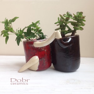 Puede incluir: Dos macetas de cerámica con forma de personas que se toman de la mano, una roja y otra marrón, con plantas verdes creciendo en su interior.