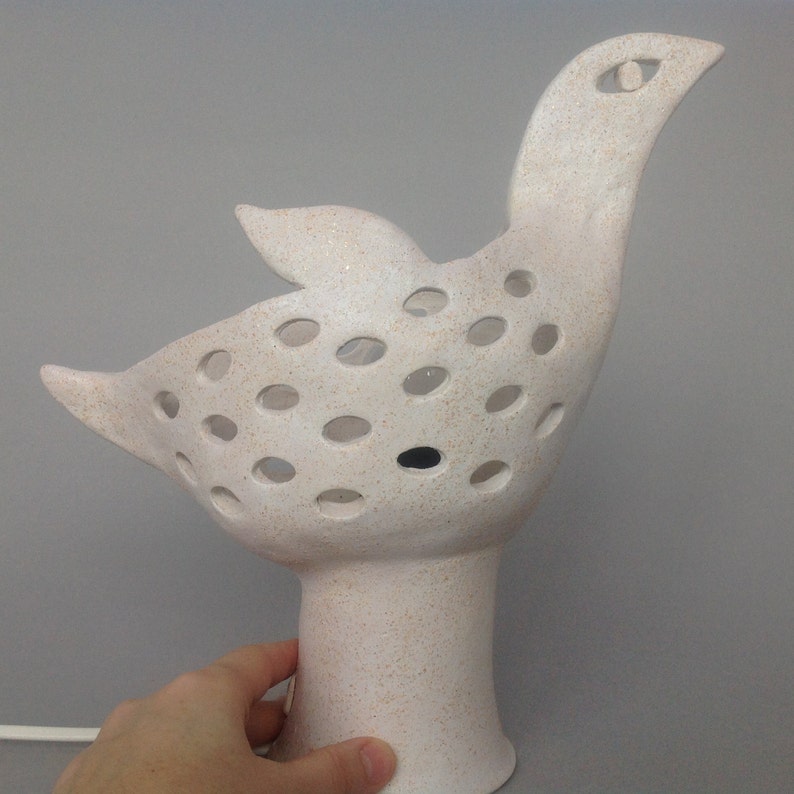 Op de afbeelding: Witte keramische vogellamp met een getextureerd oppervlak en een patroon van gaten. De lamp heeft een lange nek en een rond lichaam. De voet van de lamp is rond en heeft een getextureerd oppervlak.