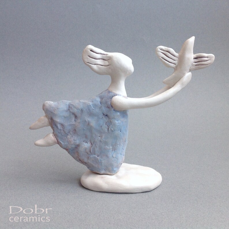 May include: A white ceramic sculpture of a woman with a blue dress, holding a white bird in her outstretched hand. The woman is in a flying pose, with her legs extended and her arms raised. The sculpture is on a white base.