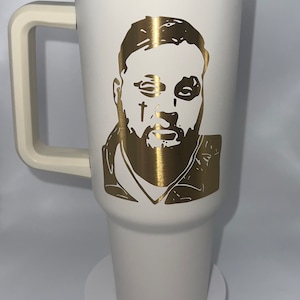 May include: White travel mug with a gold lid and handle. The mug has a gold silhouette of a man's face with a beard and a cross on his cheek.