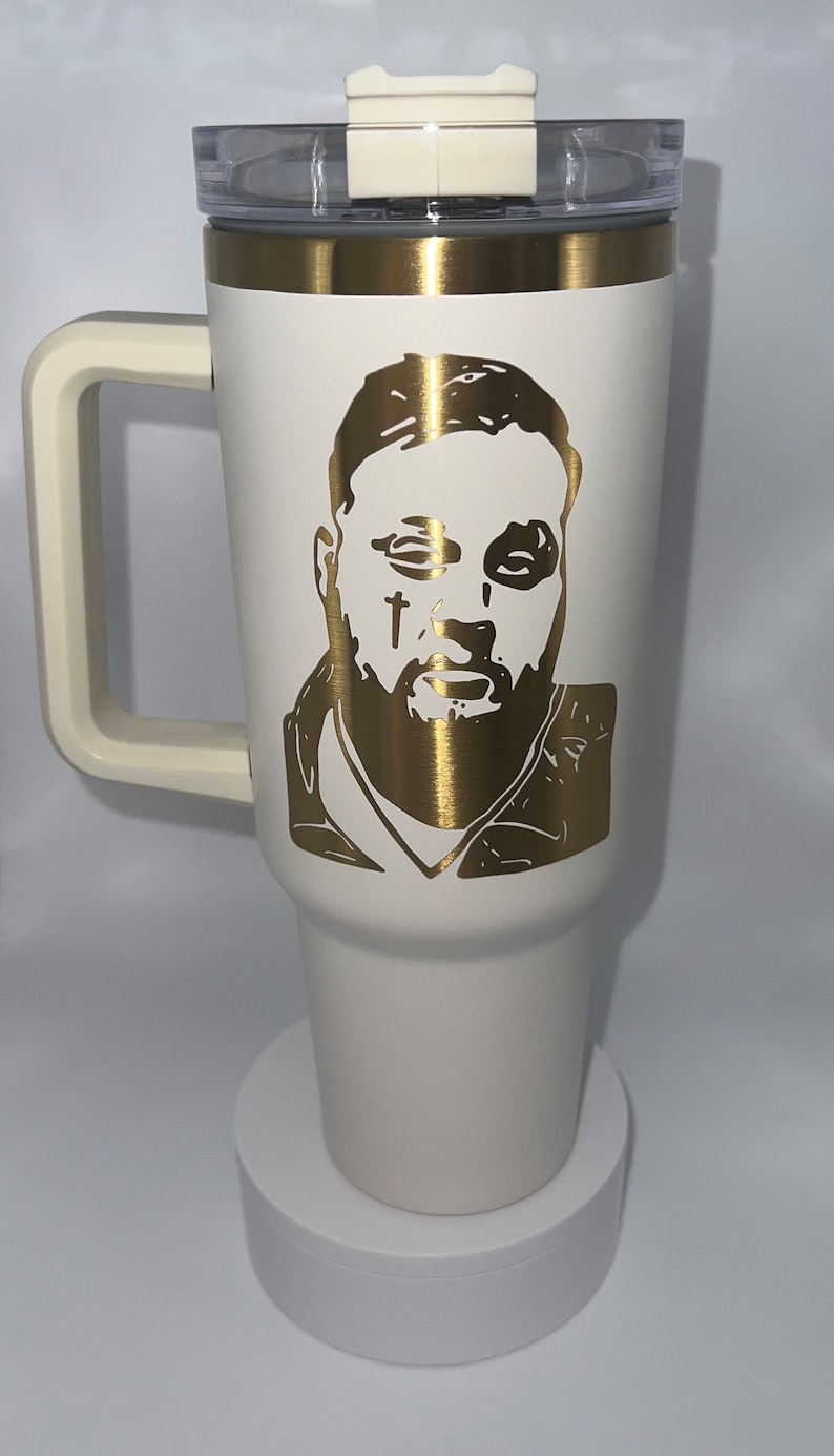 May include: White travel mug with a gold lid and handle. The mug has a gold silhouette of a man's face with a beard and a cross on his cheek.