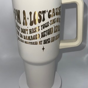 May include: White travel mug with a gold lid and handle. The mug has gold lettering that reads "I'm a lost cause baby don't waste your time on me I'm so damaged beyond repair life has shattered my hopes and dreams".