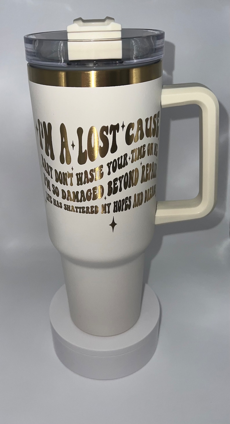 May include: White travel mug with a gold lid and handle. The mug has gold lettering that reads "I'm a lost cause baby don't waste your time on me I'm so damaged beyond repair life has shattered my hopes and dreams".