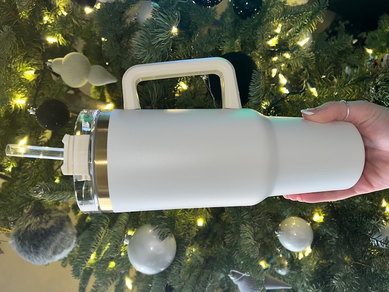 May include: White stainless steel travel mug with a handle and a clear lid. The mug has a gold accent around the top.