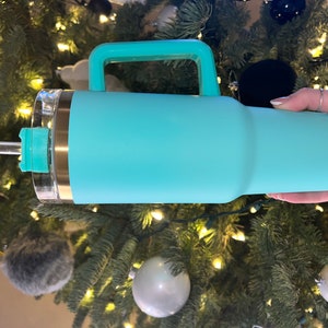 May include: A turquoise stainless steel travel mug with a handle and a clear lid. The mug has a gold band around the top and a clear straw.