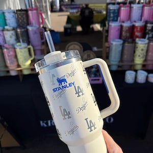 May include: Cream Stanley tumbler with the Los Angeles Dodgers logo and text. It has a handle, clear lid, and a reusable straw. Other tumblers in various colours are visible in the background.