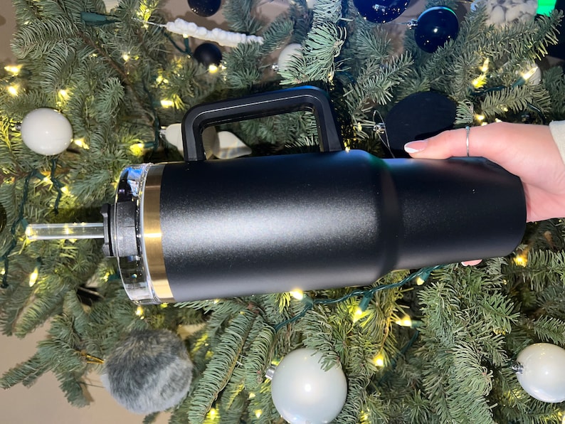 May include: A black stainless steel travel mug with a gold accent and a handle. The mug has a clear lid with a straw.