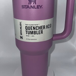 May include: A purple Stanley Quencher H2.0 Tumbler with a white lid and a black bear logo. The tumbler is 40 ounces or 1.18 liters and says "All Day Hydration".
