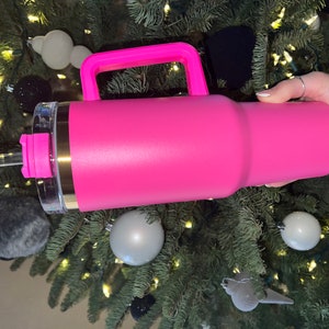 May include: A pink stainless steel travel mug with a handle and a clear lid. The mug has a gold band around the top and a straw.