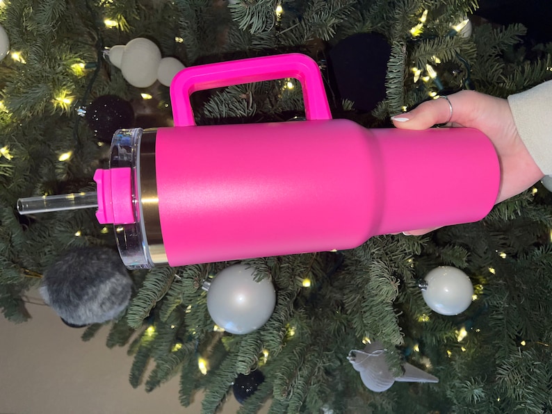 May include: A pink stainless steel travel mug with a handle and a clear lid. The mug has a gold band around the top and a straw.