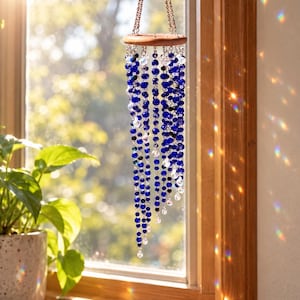 May include: A hanging mobile featuring a circular wooden top with multiple strands of deep blue and clear faceted beads suspended by copper-colored chains. The beads catch the sunlight, creating small rainbow light reflections.