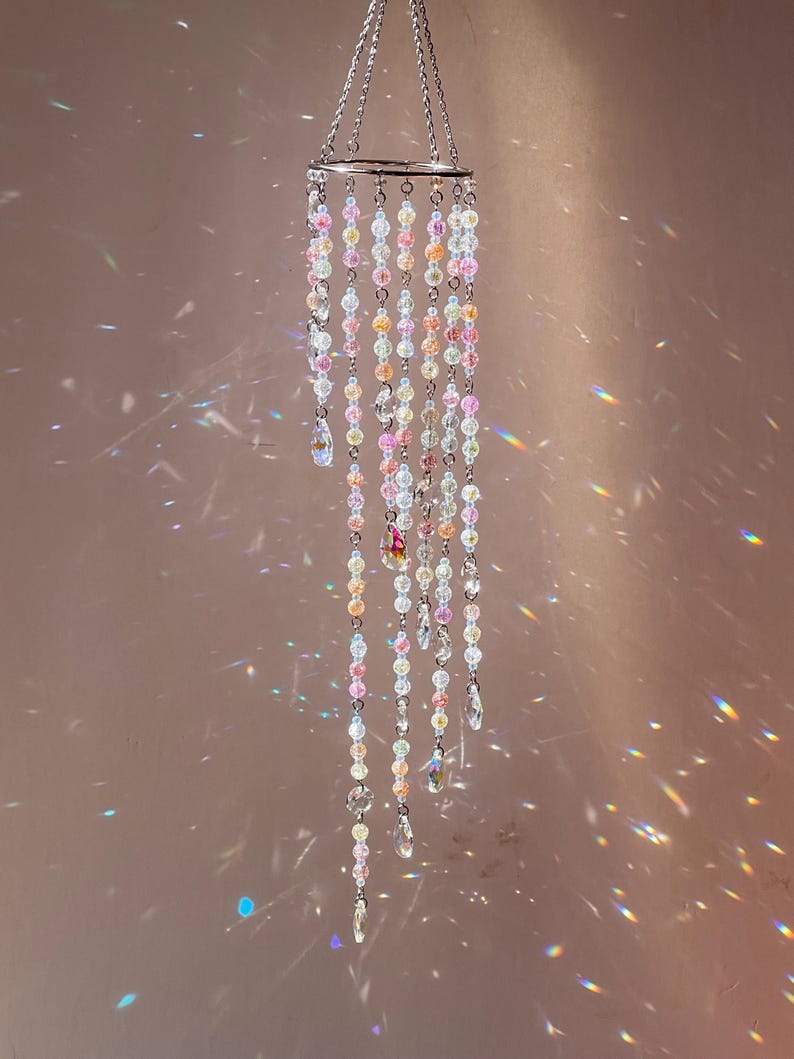 May include: A hanging mobile featuring a circular silver metal frame with multiple cascading strands of translucent beads in shades of pink, yellow, and clear. Each strand terminates in a faceted teardrop-shaped crystal pendant.