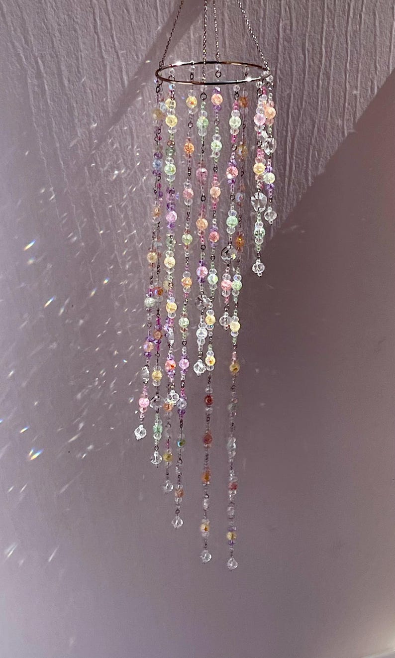 May include: A hanging mobile featuring a circular silver-toned metal frame with multiple cascading strands of translucent, multi-colored beads in shades of pink, yellow, green, and clear crystal, creating light refractions on the surrounding area.